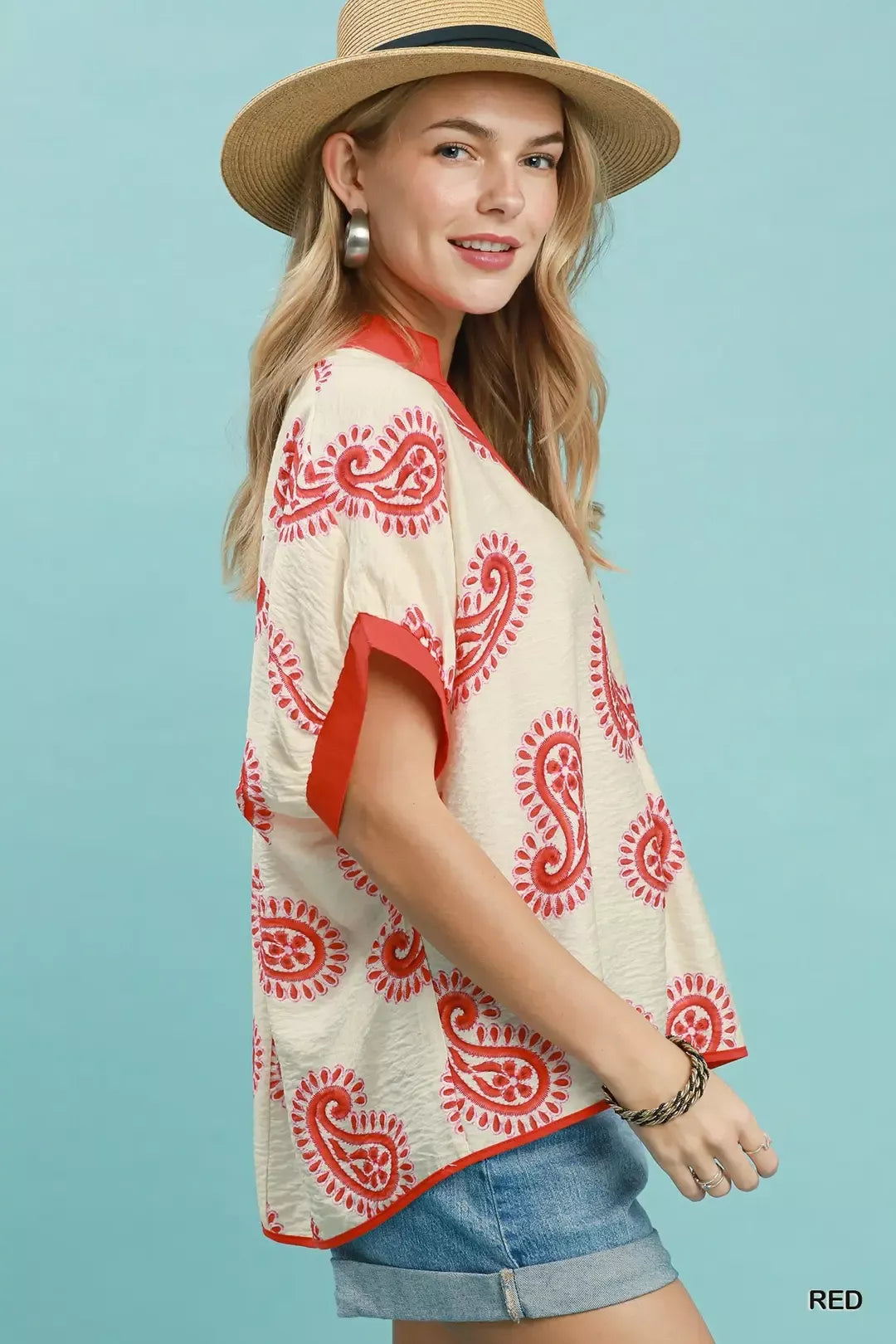 Woman wearing a cream blouse with red patterns and denim shorts against a light blue background