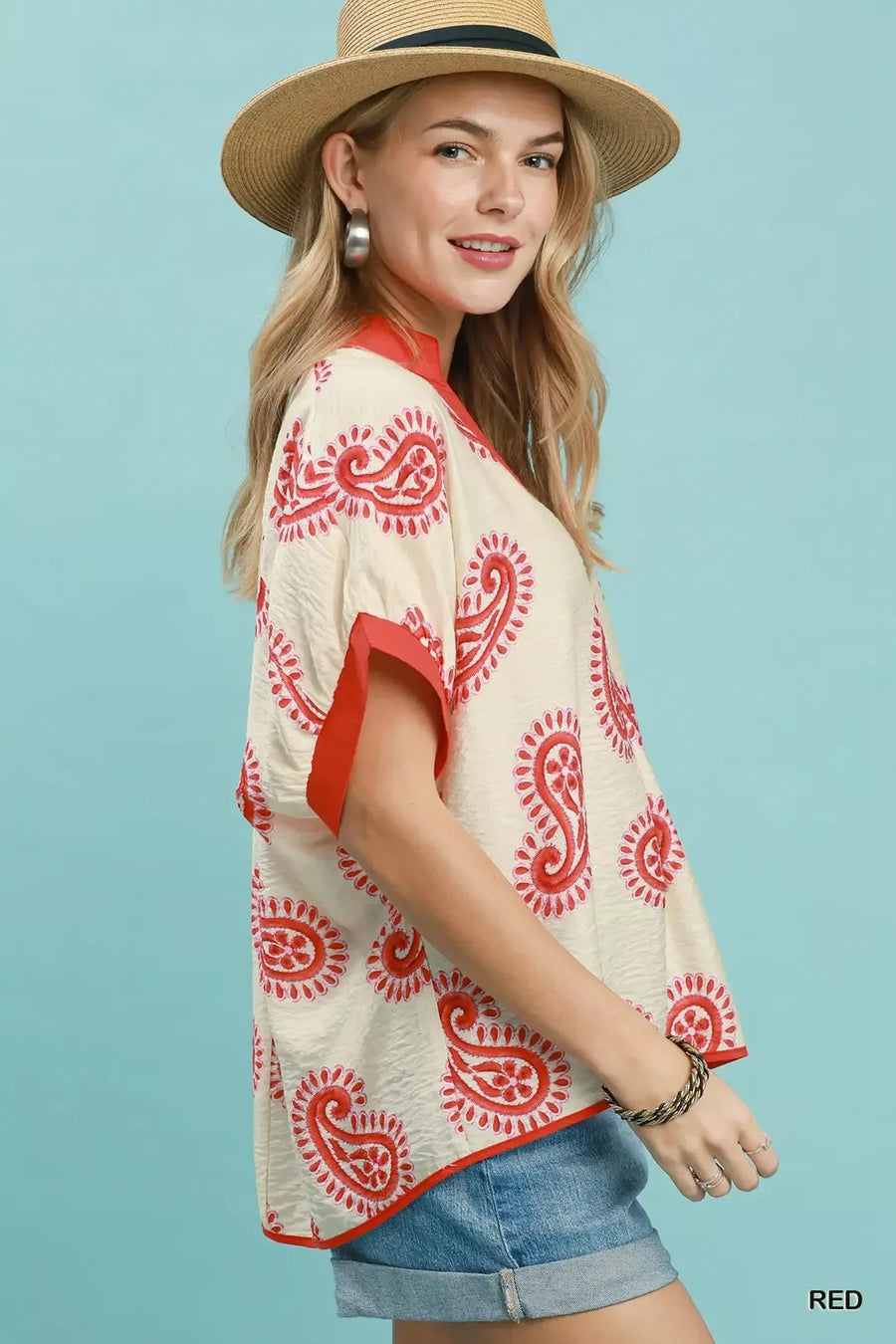 Woman wearing a cream blouse with red patterns and denim shorts against a light blue background