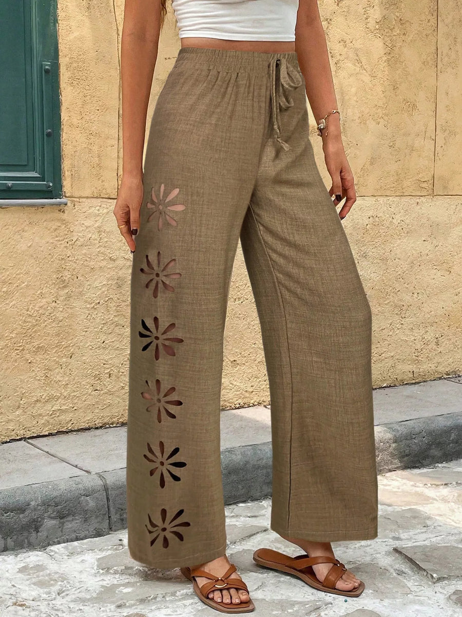 Person wearing brown wide-leg pants with floral cutouts against a beige wall.