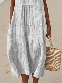 Person wearing a light gray dress holding a woven bag on a beach.