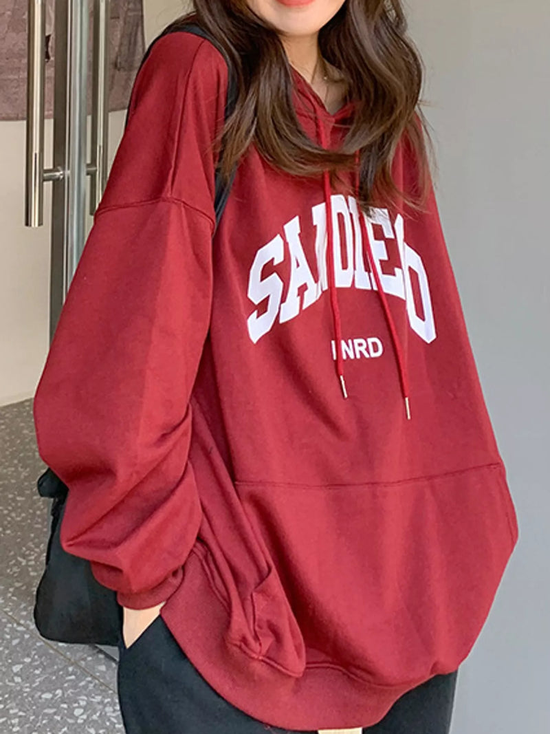A woman wearing an oversized maroon hoodie with white collegiate-style lettering that reads 'SAIDED' and smaller text 'INRD' underneath, paired with black pants and a black shoulder bag.