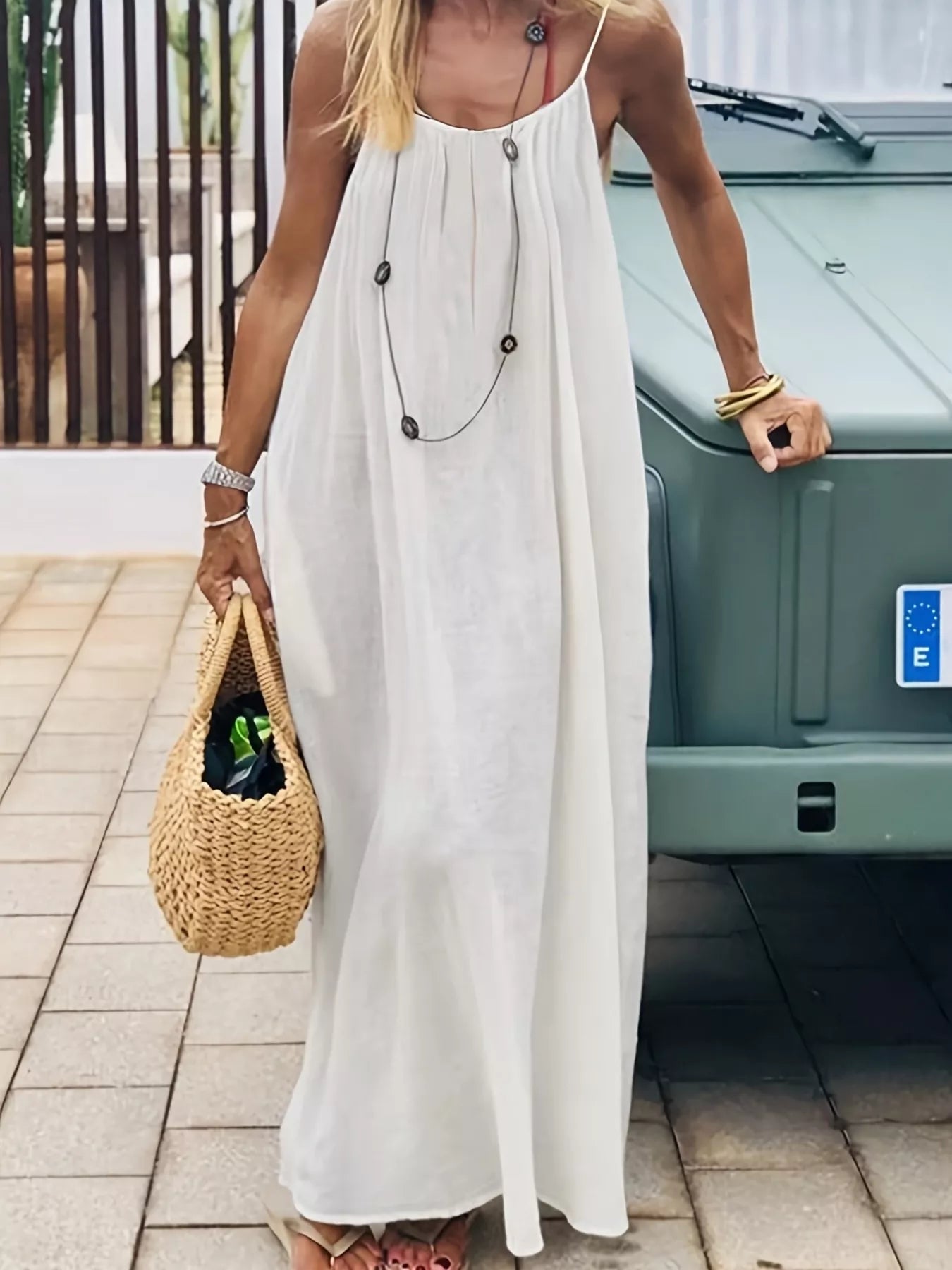 Woman in a white dress holding a woven bag, standing next to a green vehicle.