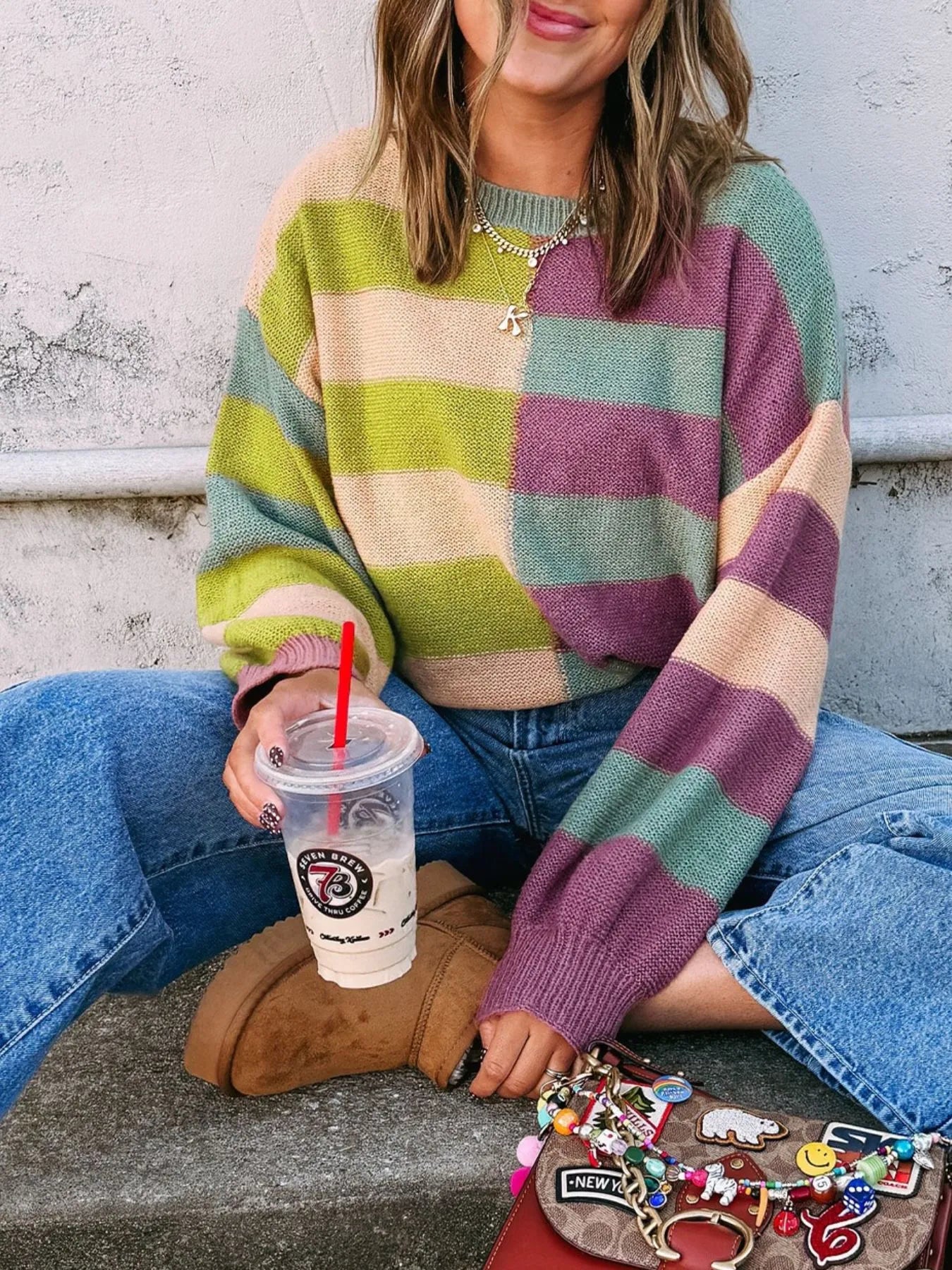 Person wearing a colorful striped sweater sitting on a concrete surface with a drink.