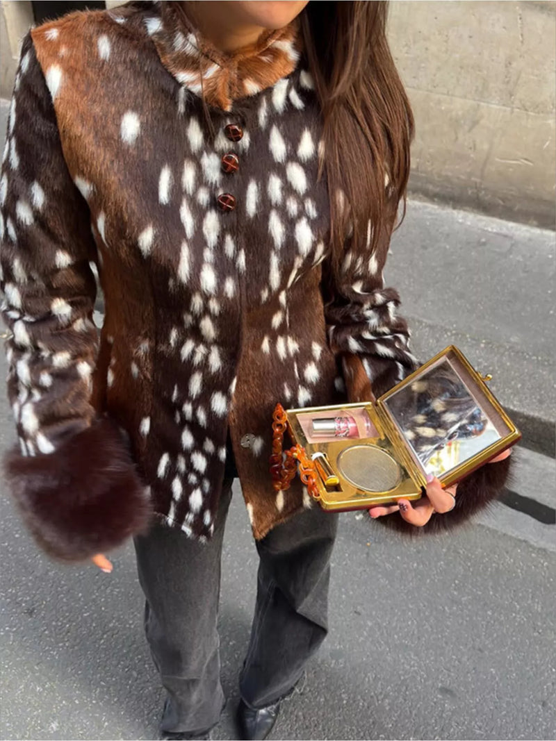 A person wearing a brown and white spotted faux fur jacket with dark gray pants, holding an open gold-framed compact mirror in an outdoor street setting.