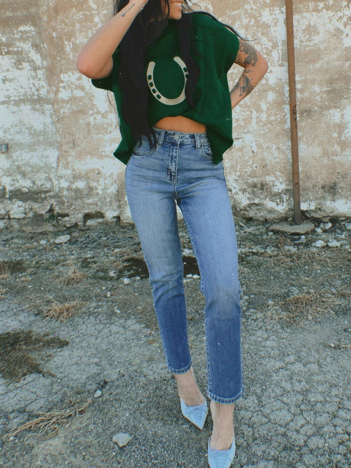 Person wearing a green crop top with a horse shoe design and blue jeans against a textured wall.