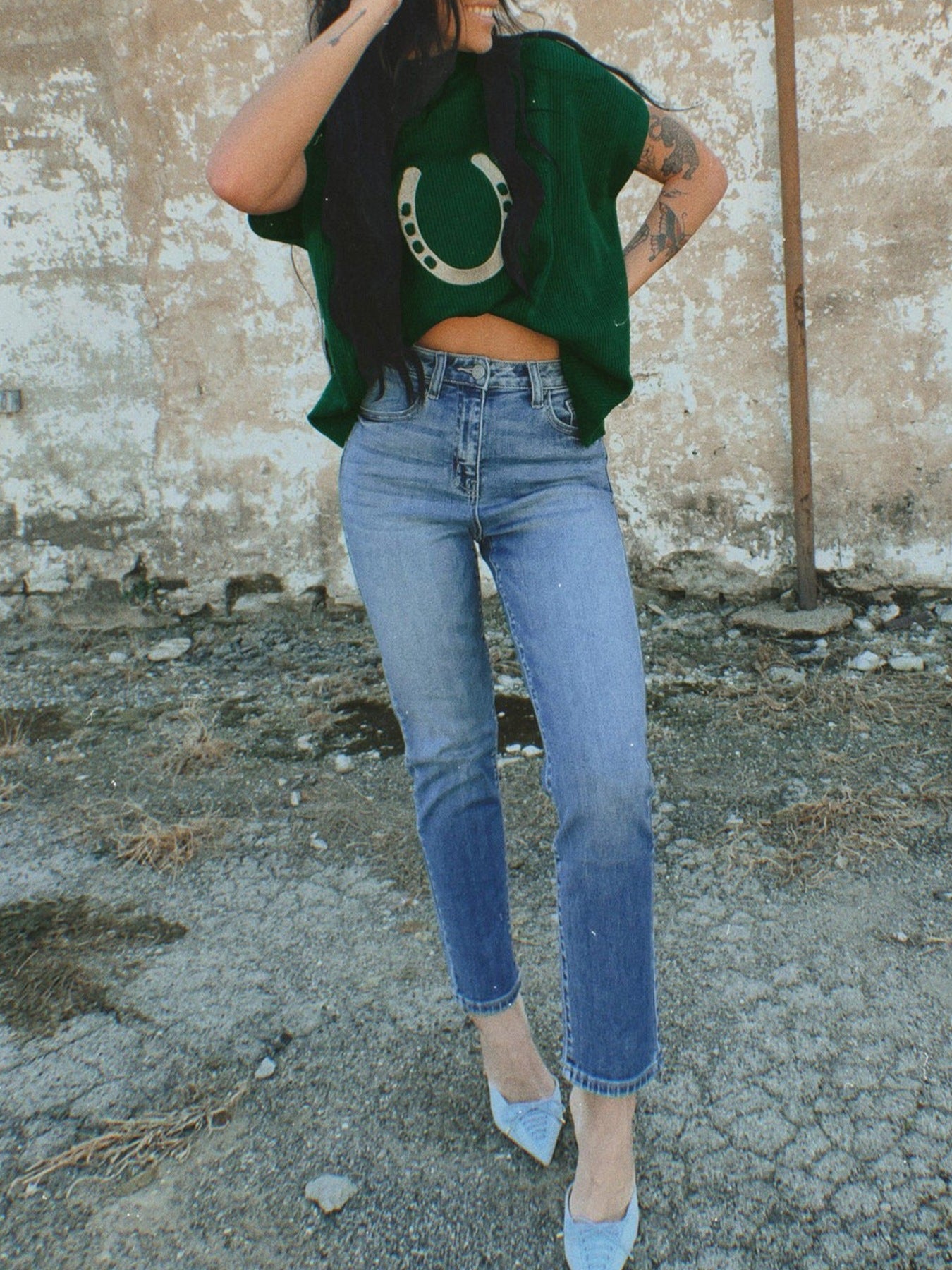 Person wearing a green crop top with a horse shoe design and blue jeans against a textured wall.