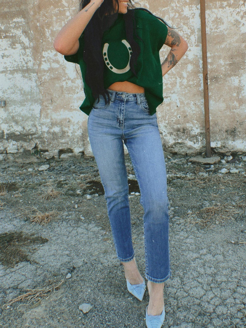 Person wearing a green crop top with a horse shoe design and blue jeans against a textured wall.