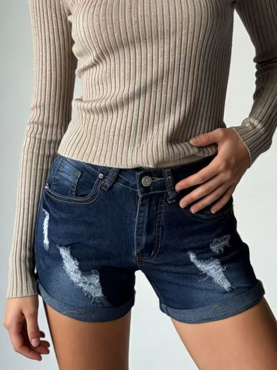 Person wearing a beige ribbed sweater and distressed blue denim shorts on a gray background