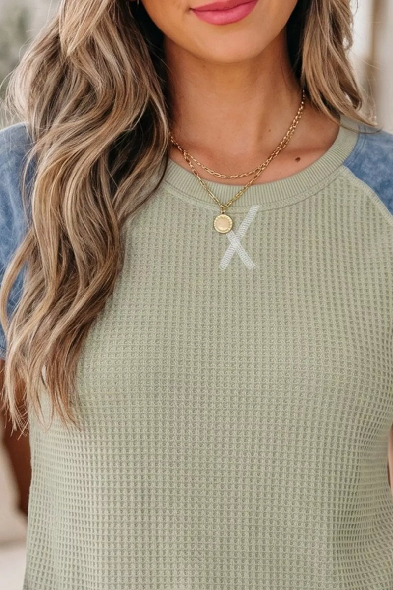 Woman wearing a green top with blue sleeves and layered gold necklaces.