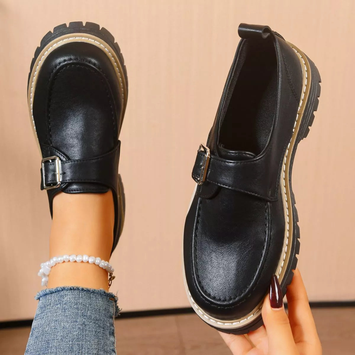 Black leather shoes with a strap held by a person against a beige background