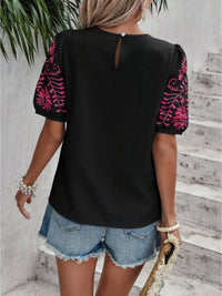 Person wearing a black top with pink floral sleeves and denim shorts against a light gray wall.