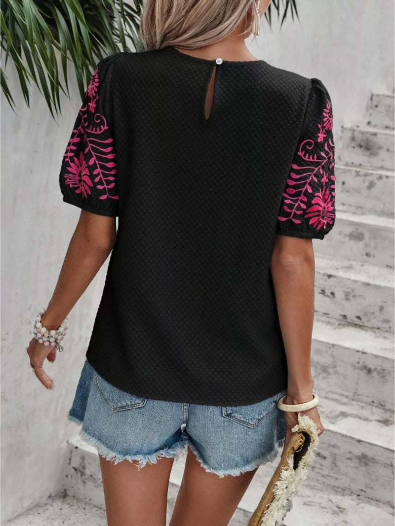 Person wearing a black top with pink floral sleeves and denim shorts against a light gray wall.