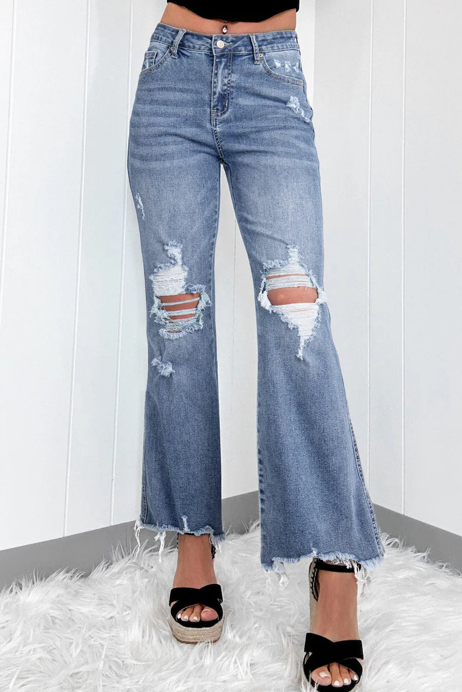 A pair of distressed, raw hem bootcut jeans with a slightly stretchy fabric, displayed on a person against a white background with a furry white surface.