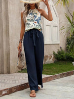 Woman wearing a floral top and navy pants with a straw hat and bag, standing on a stone path.