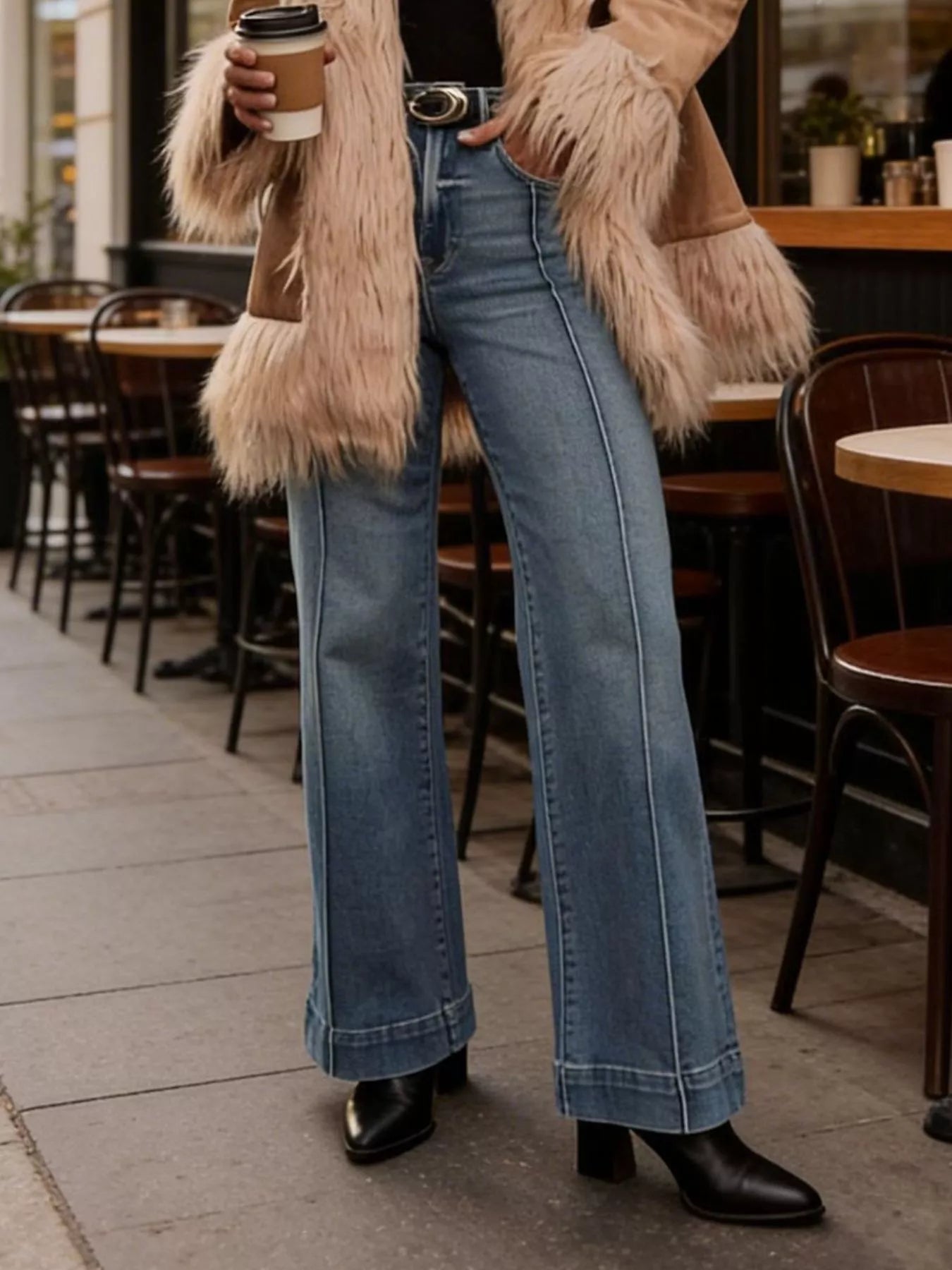 Person wearing a fur coat, blue jeans, and black shoes standing outdoors near a cafe.