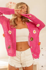 Woman wearing a pink crochet cardigan with floral decorations, white top, and shorts indoors.
