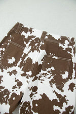 Close-up of the back waistband and pockets of brown and white cow-print denim jeans on a light gray surface.