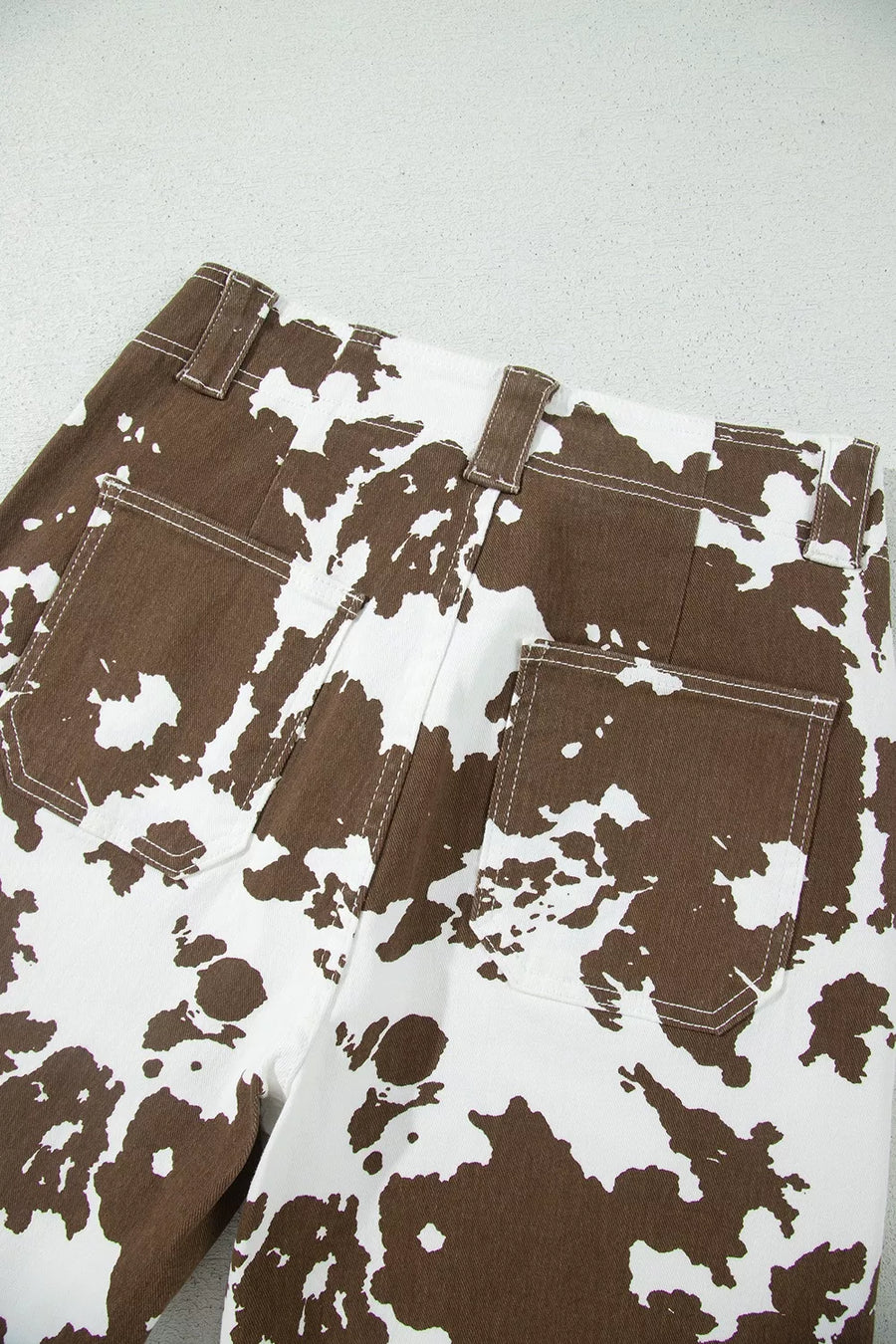 Close-up of the back waistband and pockets of brown and white cow-print denim jeans on a light gray surface.