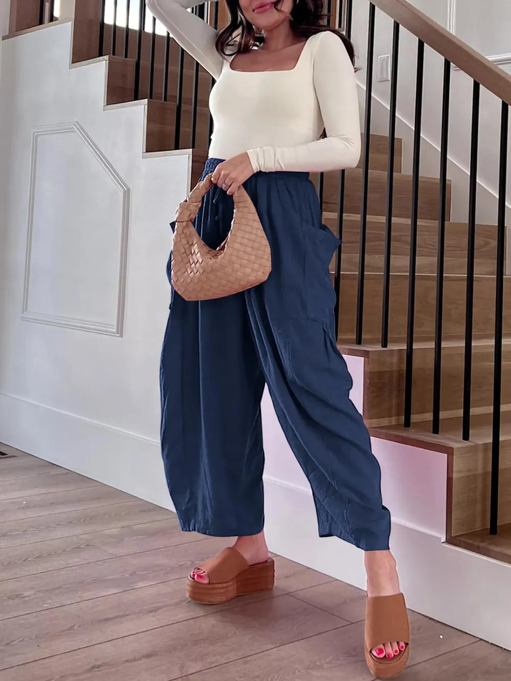 Woman in a white top and blue pants holding a woven handbag on a wooden floor.