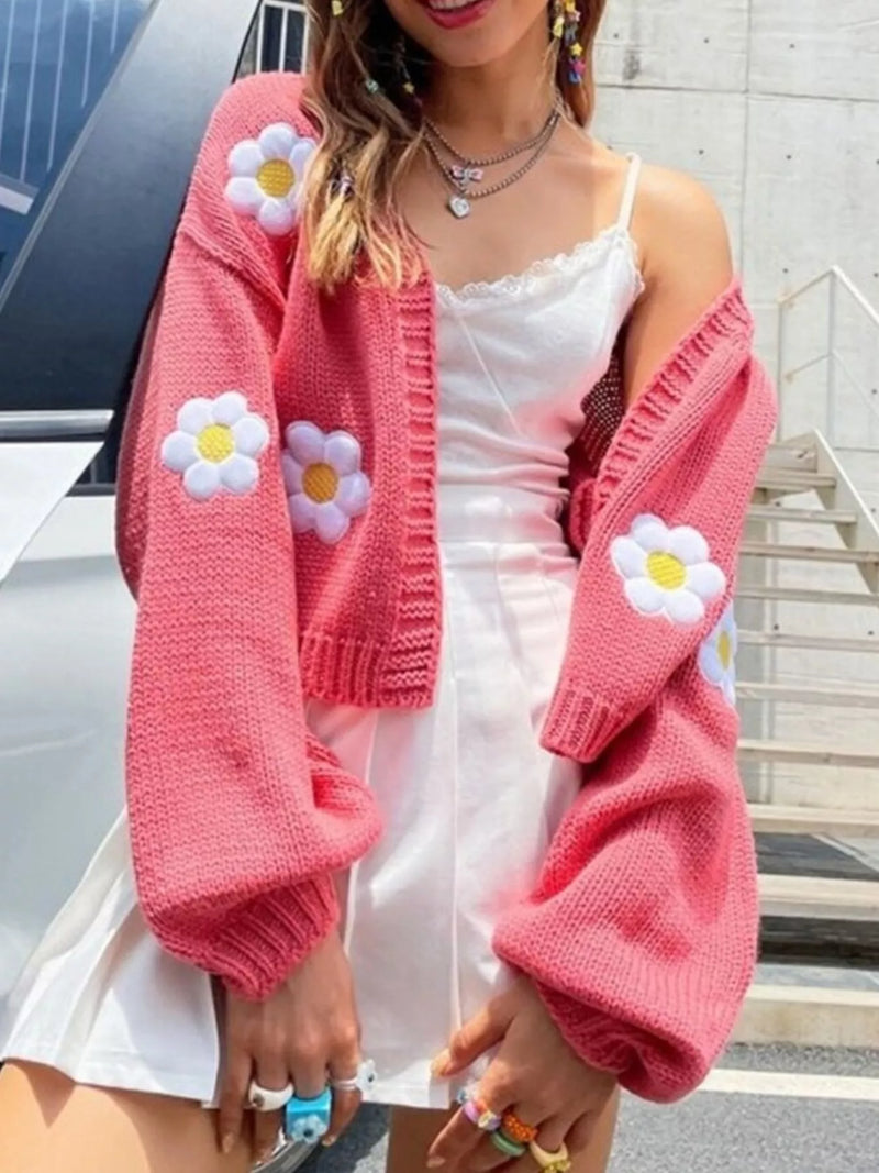 A woman wearing a pink knit cardigan with large white daisy patches, styled over a white camisole dress in an outdoor urban setting.