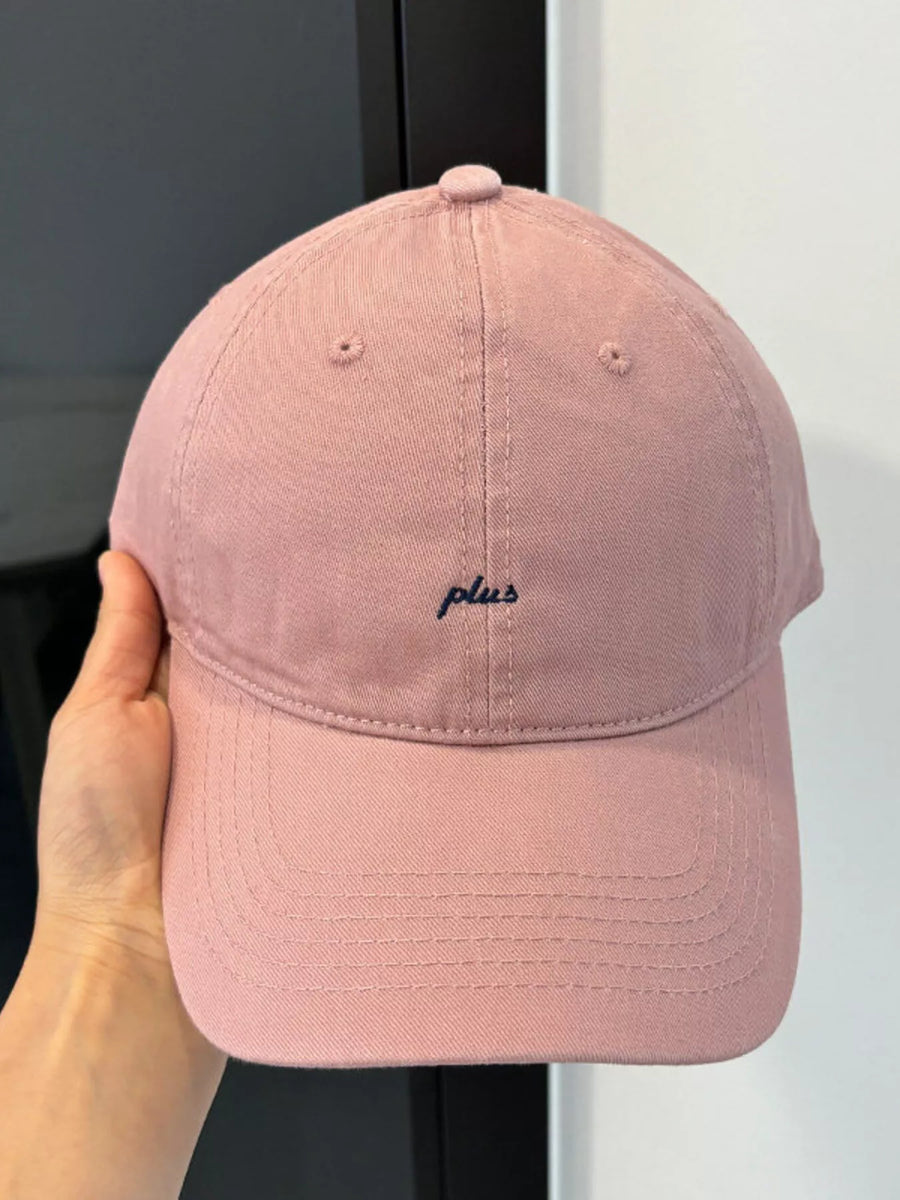 Pink cap with 'plus' logo held by a hand against a neutral background