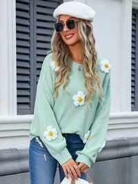 Woman wearing a light green sweater with white daisy flowers, blue jeans, and a white beret.