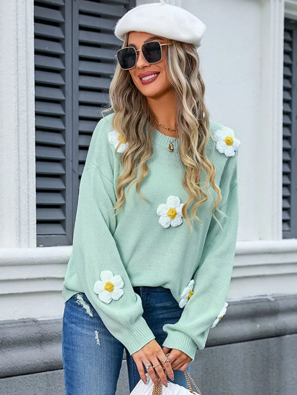 Woman wearing a light green sweater with white daisy flowers, blue jeans, and a white beret.