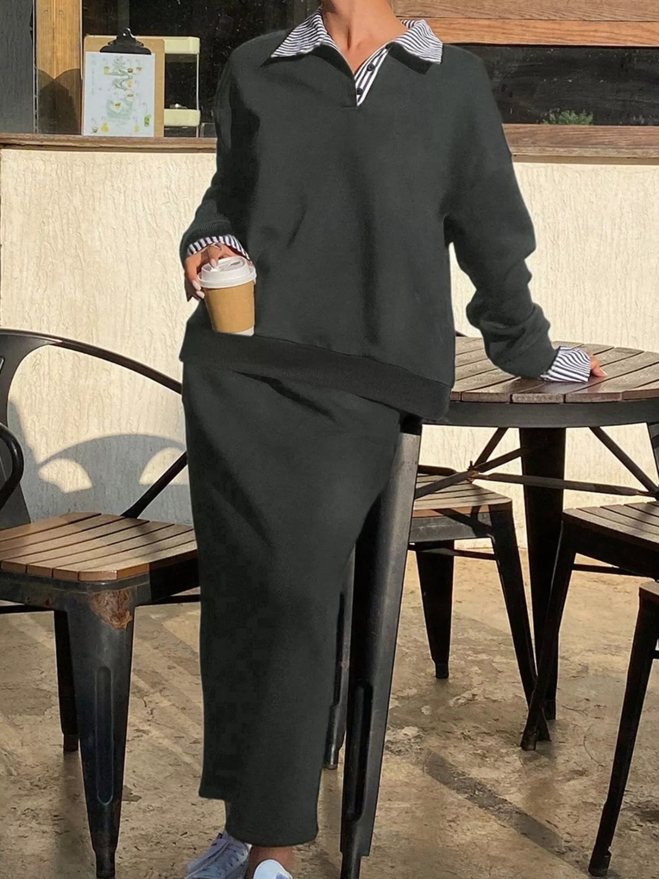 Person wearing a dark outfit with striped shirt collar, holding a coffee cup outdoors.