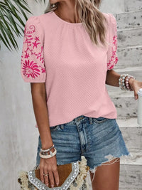 Person wearing a pink top with floral embroidery and denim shorts, holding a white cup.