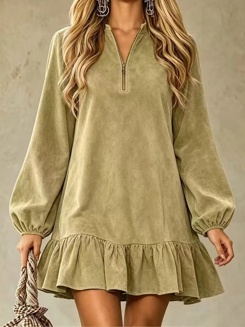 A woman wearing a sage green long-sleeve mini dress with a half-zip neckline and a ruffled hem, holding a checkered bag against a textured gray background.