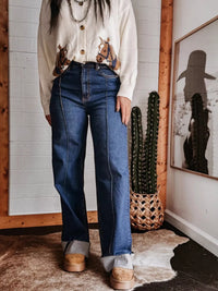 Person wearing a patterned blouse and blue jeans standing on a wooden floor with a white wall background