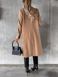 Person wearing a beige coat holding a black handbag against a gray concrete wall.