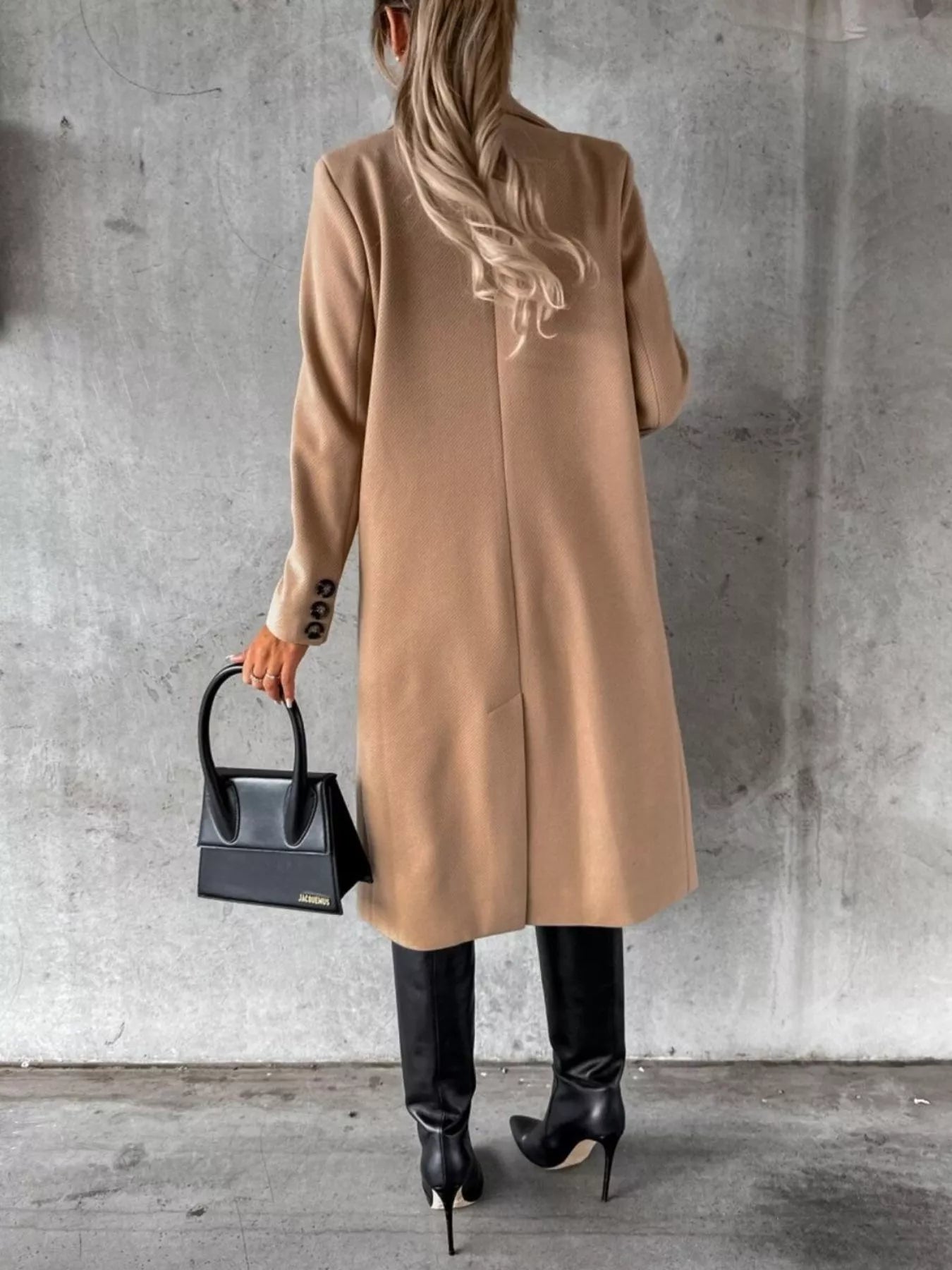 Person wearing a beige coat holding a black handbag against a gray concrete wall.