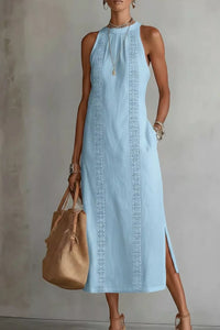 Woman wearing a light blue dress with intricate patterns, holding a beige handbag against a neutral background.