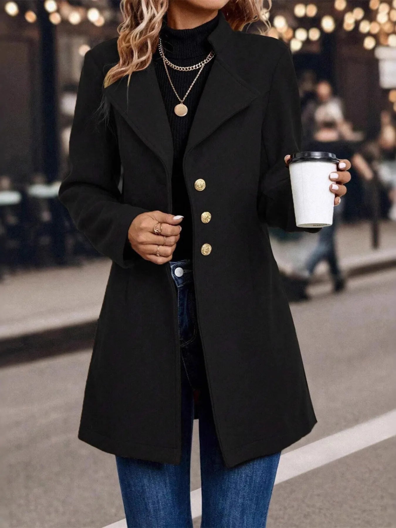 Person wearing a black coat with gold buttons, holding a coffee cup outdoors.