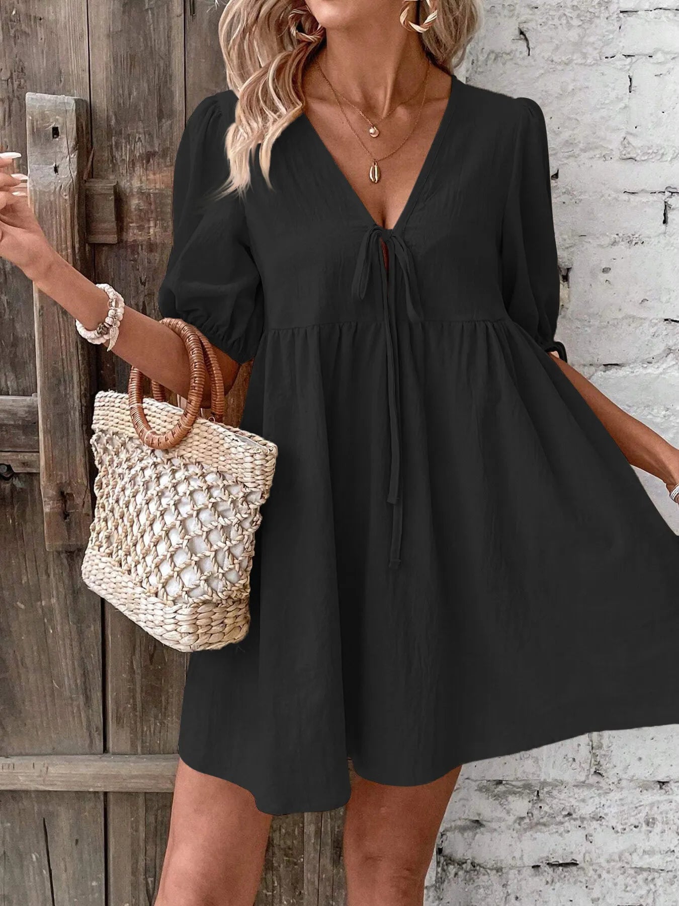 Woman wearing a black dress holding a woven bag against a rustic wooden and brick wall.