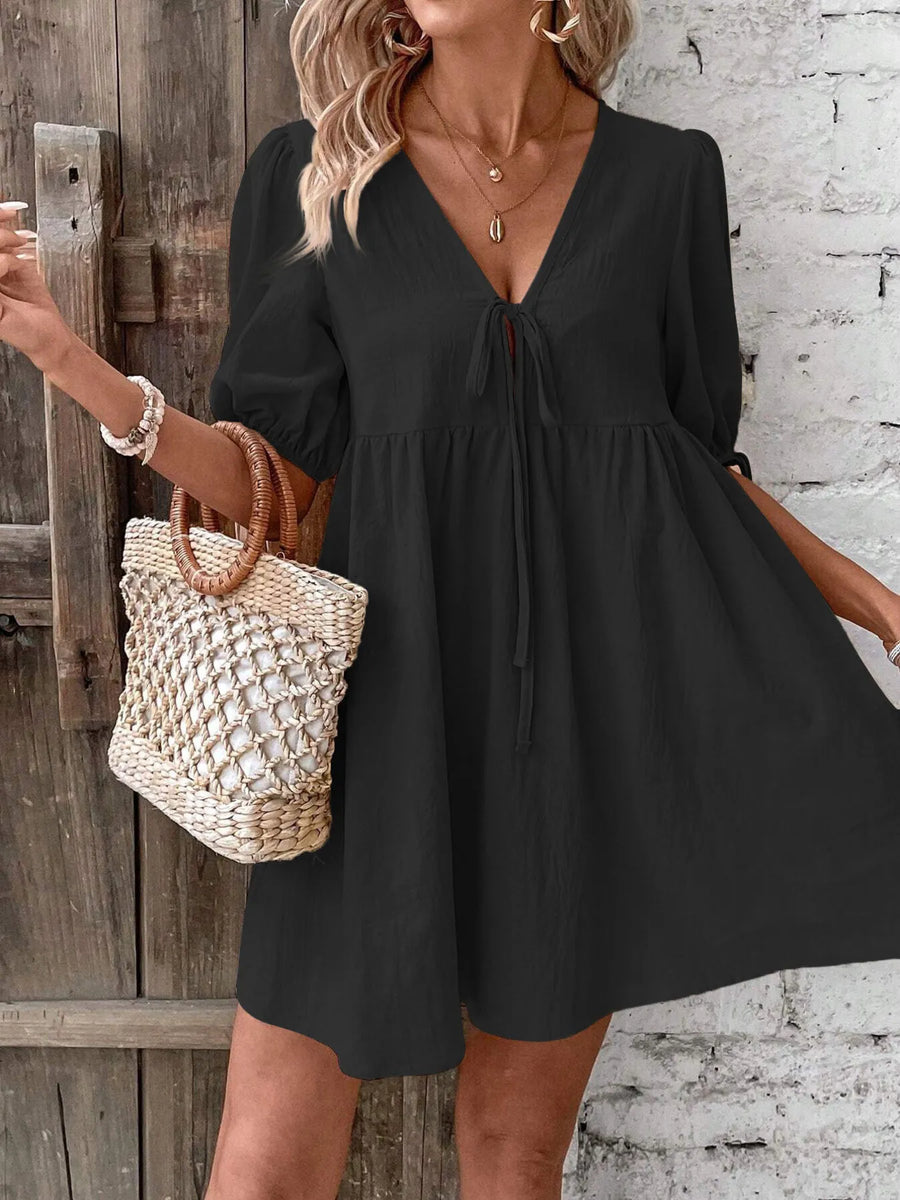 Woman wearing a black dress holding a woven bag against a rustic wooden and brick wall.