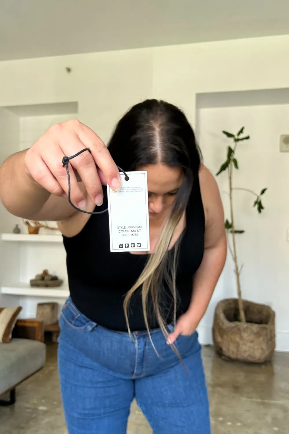 Person holding a clothing tag in a room with a plant and shelves.