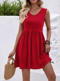 Woman wearing a red dress holding a straw bag against a white wall with greenery.