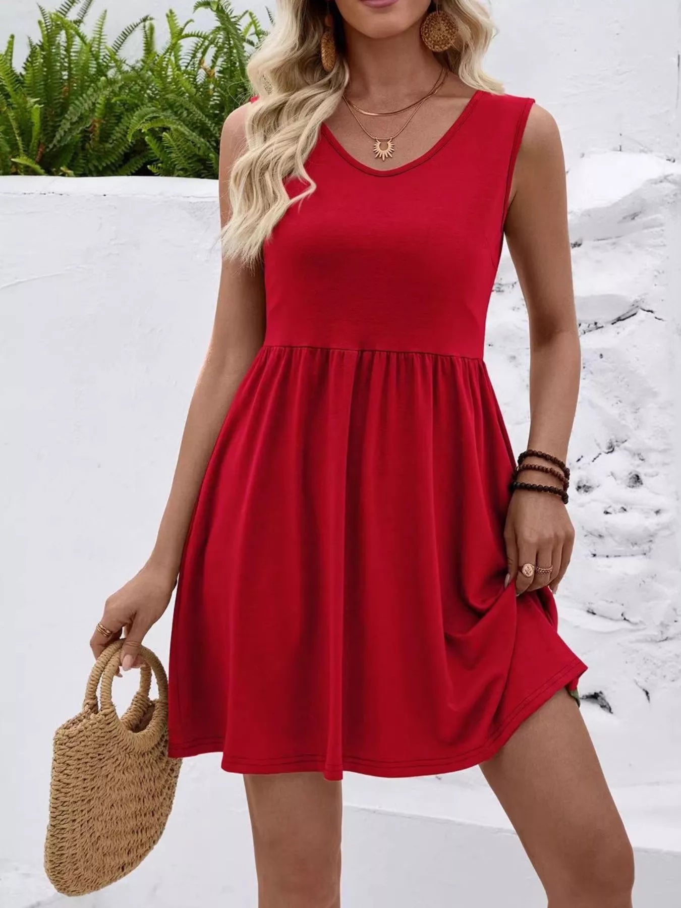 Woman wearing a red dress holding a straw bag against a white wall with greenery.