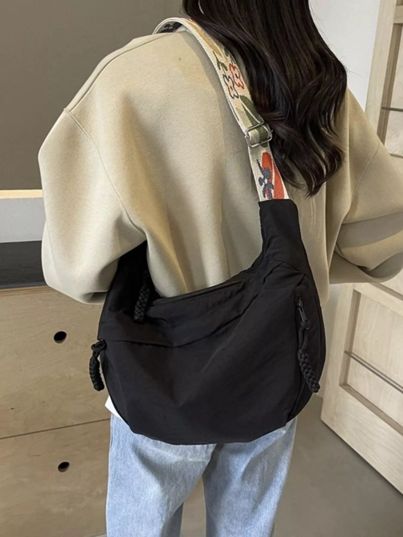 Person wearing a black crossbody bag with a beige jacket and light blue jeans indoors.