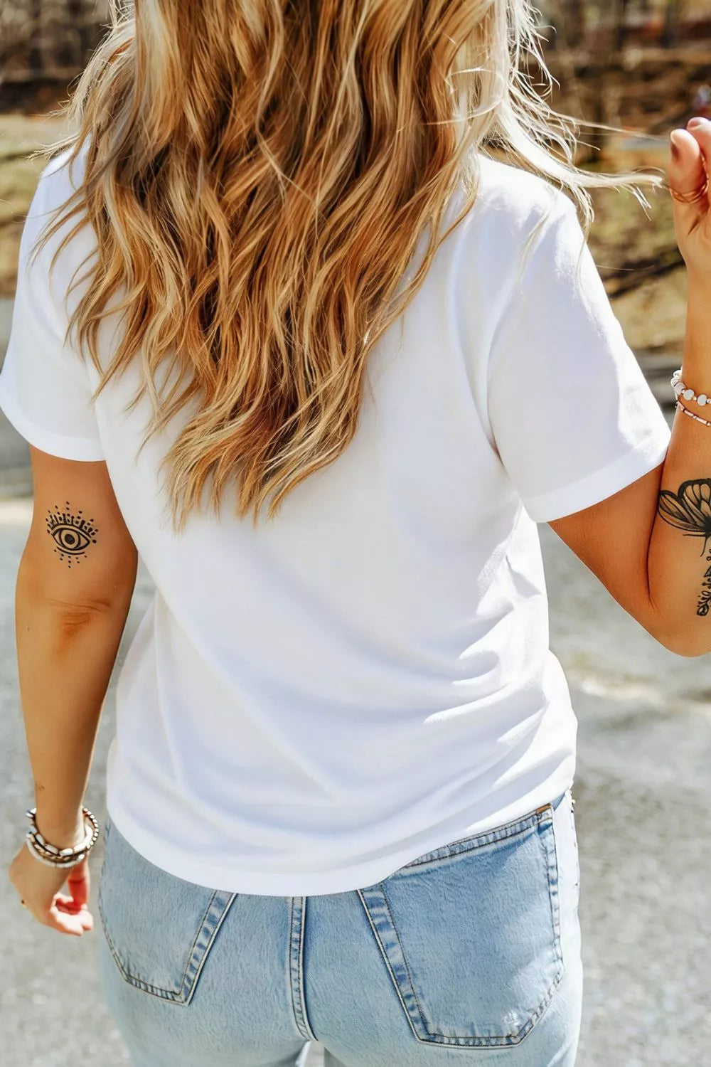 Person wearing a white t-shirt and light blue jeans with tattoos on arms, standing outdoors.