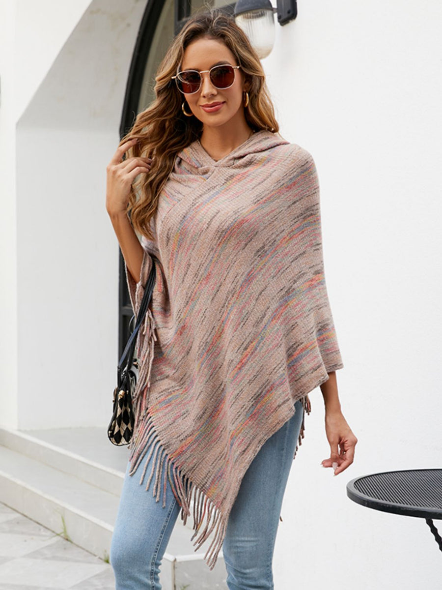 A woman wearing a hooded poncho with fringe, styled with light blue jeans, sunglasses, and a patterned handbag, standing in front of a white building.