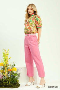 Woman wearing a colorful floral blouse and pink pants standing next to a plant on a white background
