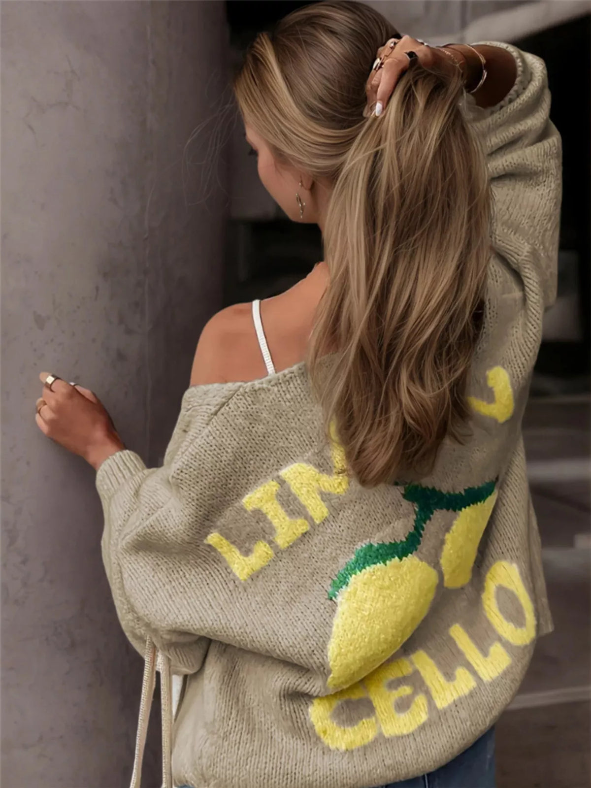 A woman wearing a beige knit sweater with a yellow and green lemon graphic and the words 'LIME CELLO' on the back, shown from behind in an indoor setting.