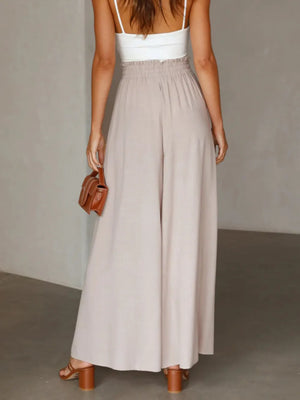 Woman wearing a long beige skirt with a white top and brown sandals, holding a brown handbag against a plain background.