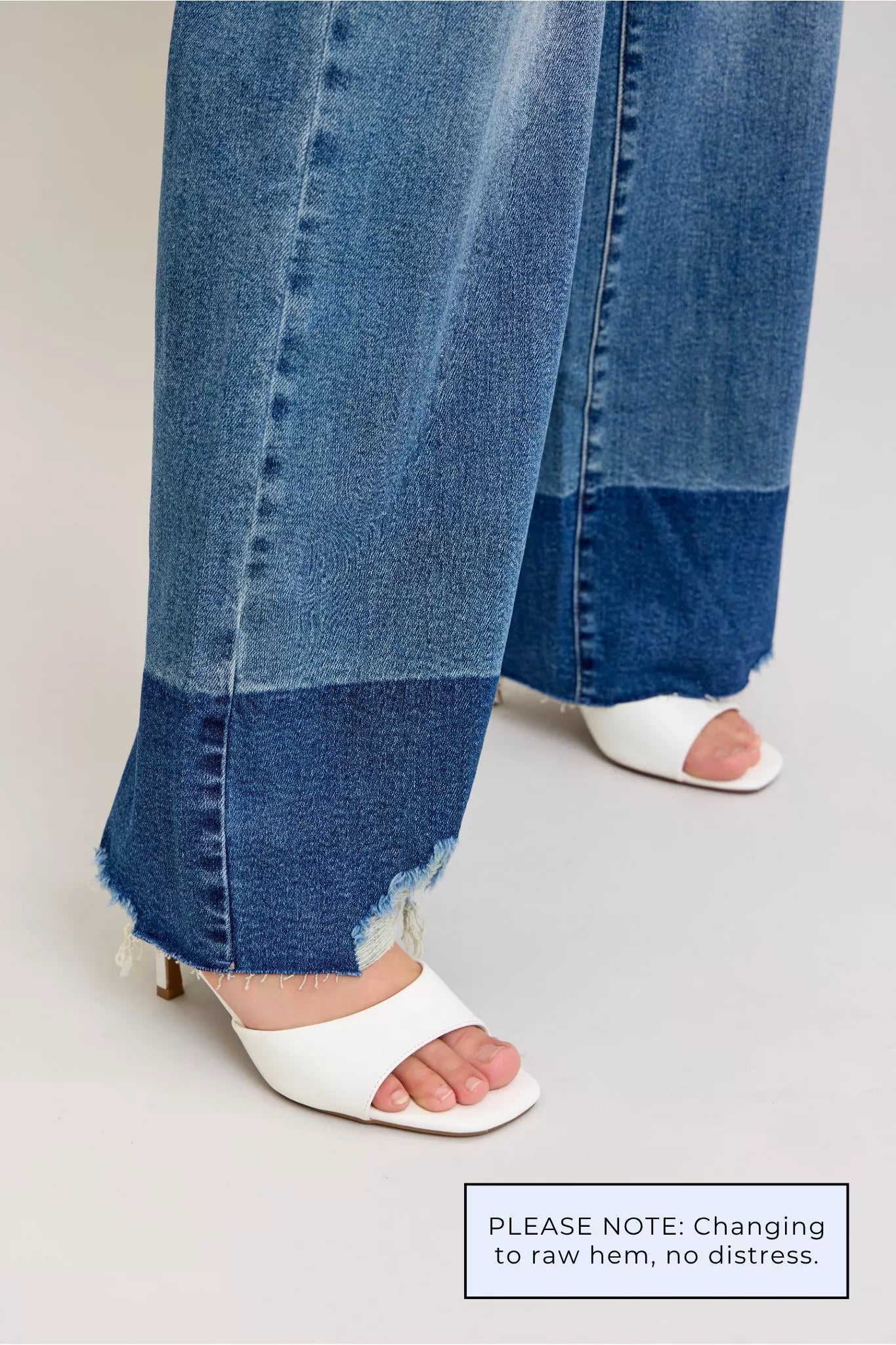 Close-up of blue jeans with a raw hem and white sandals on a light gray background.