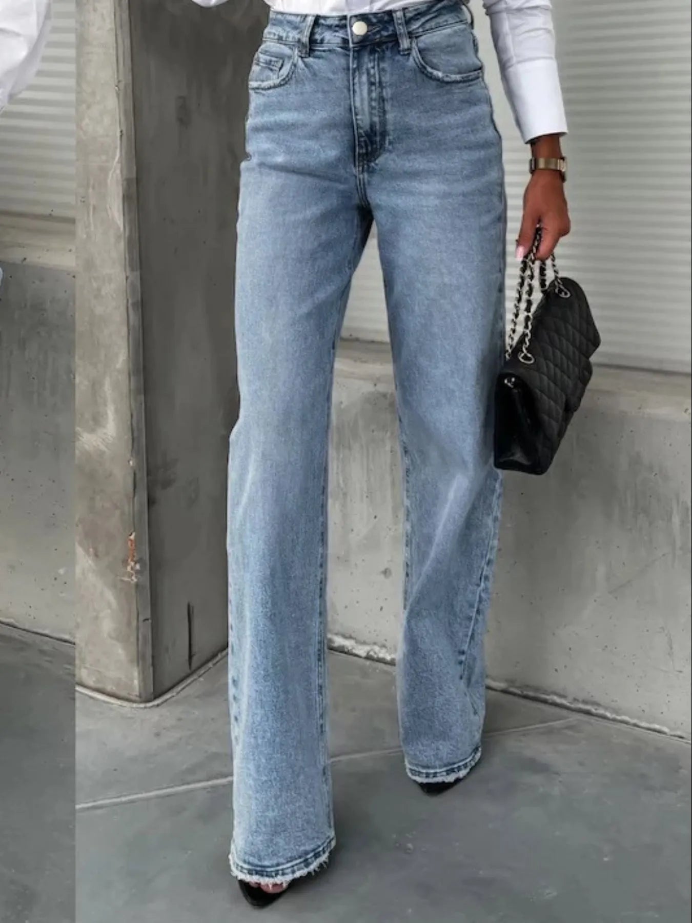 Person wearing high-waisted blue jeans holding a black handbag against a concrete wall.