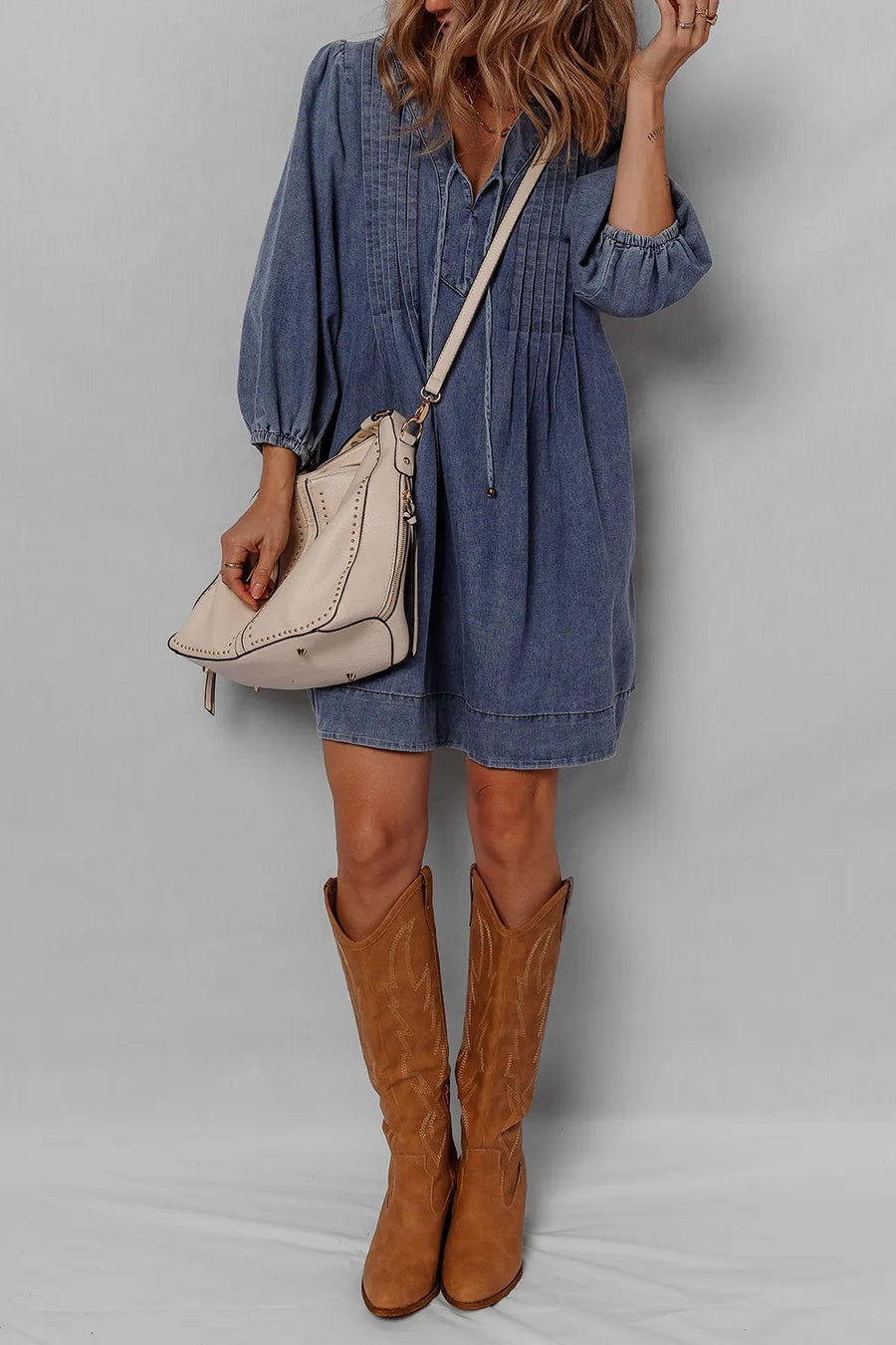 Person wearing a blue dress and brown knee-high boots with a beige handbag on a gray background