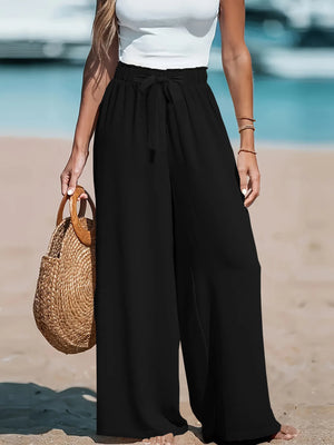 Person wearing black wide-leg pants and a white top on a beach, holding a woven bag.